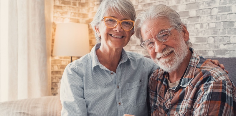 An elderly man and woman both wearing glasses are smiling in their living room.
