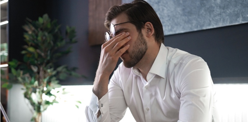 Man rubbing his eyes while sitting at a desk, looking fatigued.