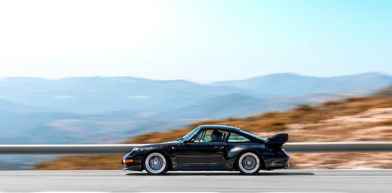 Black Porsche sports car driving on a winding mountain road, showcasing the performance heritage that inspires Porsche Design eyewear.]