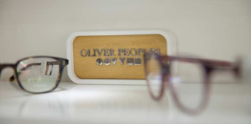 A close up of a wooden block with the text ‘OLIVER PEOPLES’ and their distinct symbols underneath. There is a pair of out of focus classes in the foreground and another pair to the left of the block.