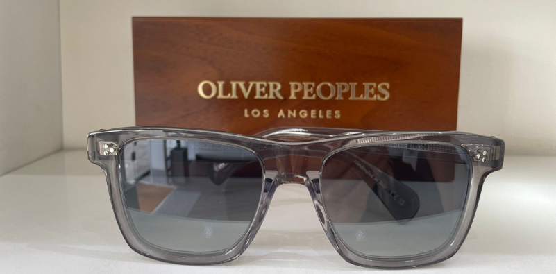 A pair of grey Oliver Peoples sunglasses in front of a wooden block that has the text ‘OLIVER PEOPLES LOS ANGELES’ in gold.