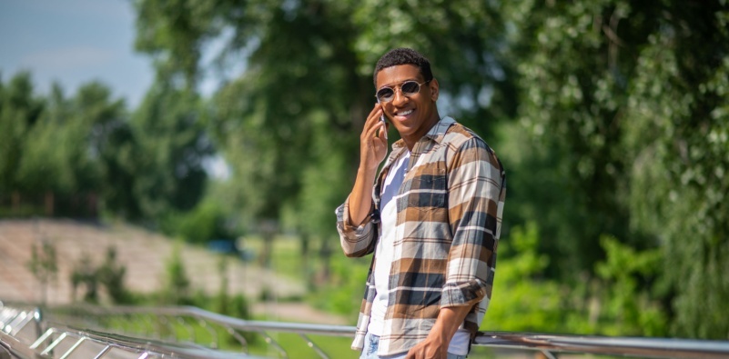 A photo of a fashionable gentleman wearing a checked shirt and metal sunglasses, on the phone outside on a sunny day.