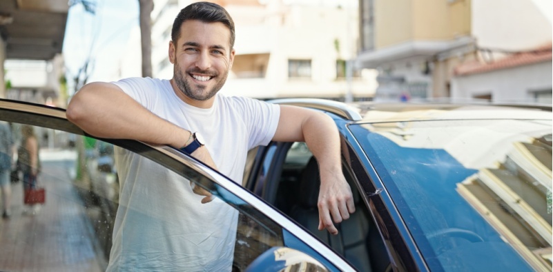 A middle aged man wearing a white t-shirt leaning of the open door of his car, smiling