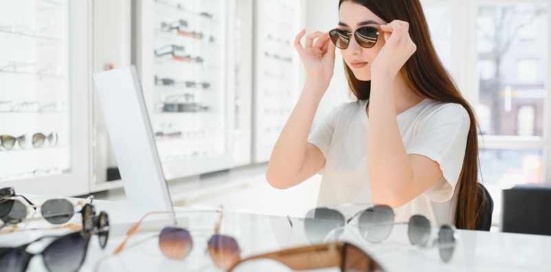 A young woman trying on sunglasses in the mirror at the opticians, with a variety of different sunglasses styles on the counter in front of her.