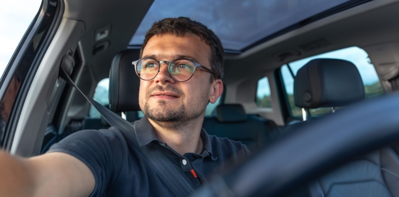 A young man sat behind the steering wheel of a car, wearing a pair of round, black glasses
