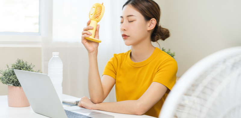 Woman using a handheld fan while working on a laptop indoors