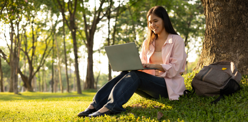 Woman working on a laptop while sitting beneath a tree in a park